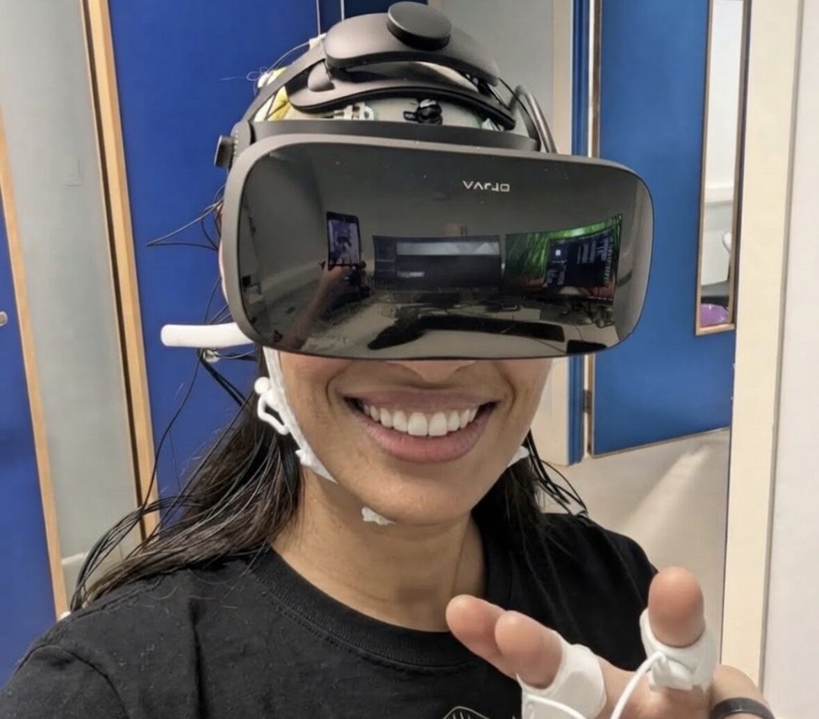 Ashlei Lewis wearing a VR headset over an EEG cap, smiling and gesturing toward the camera.