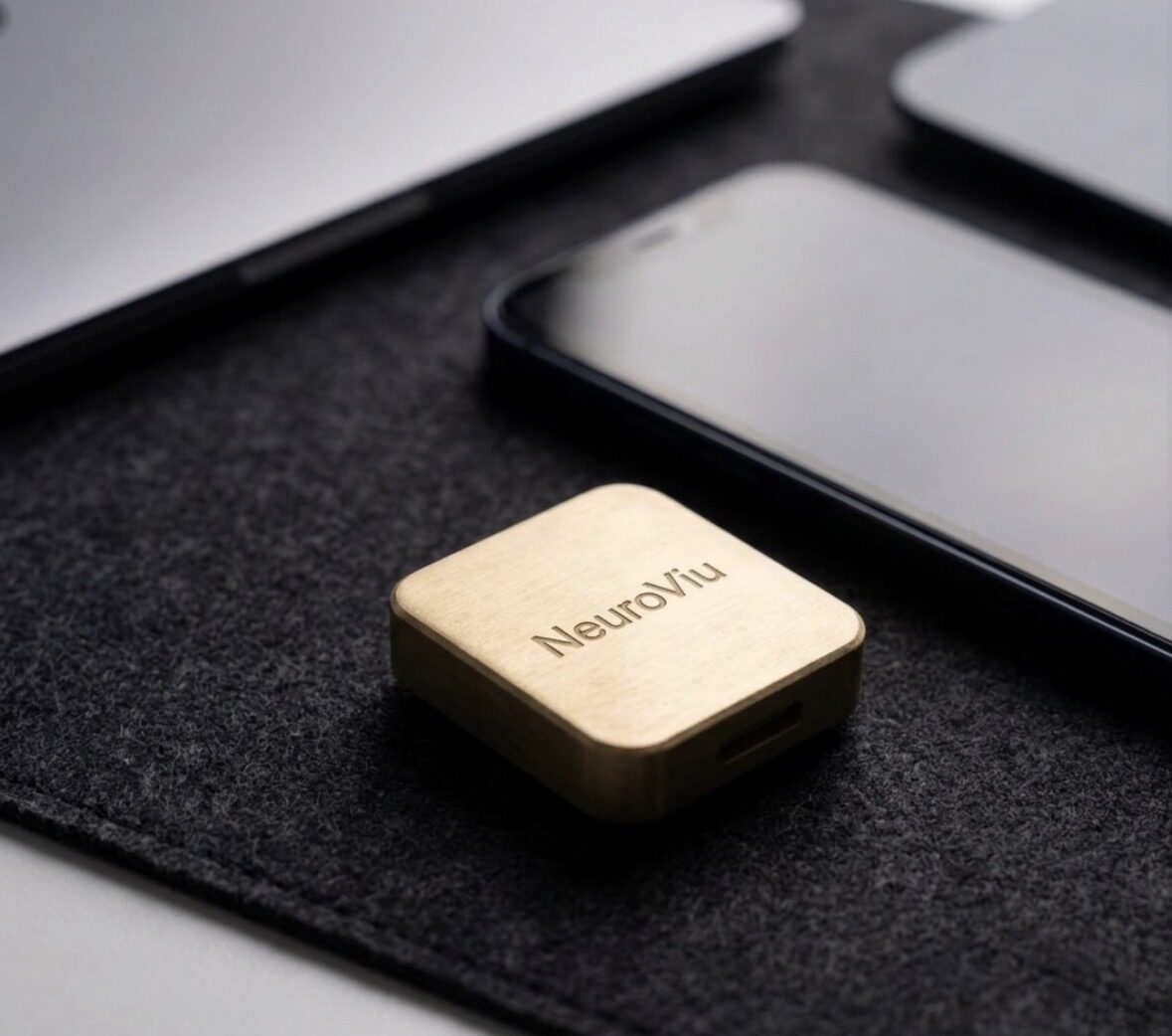 The gold NeuroViu module on a felt desk mat beside a laptop and phone.