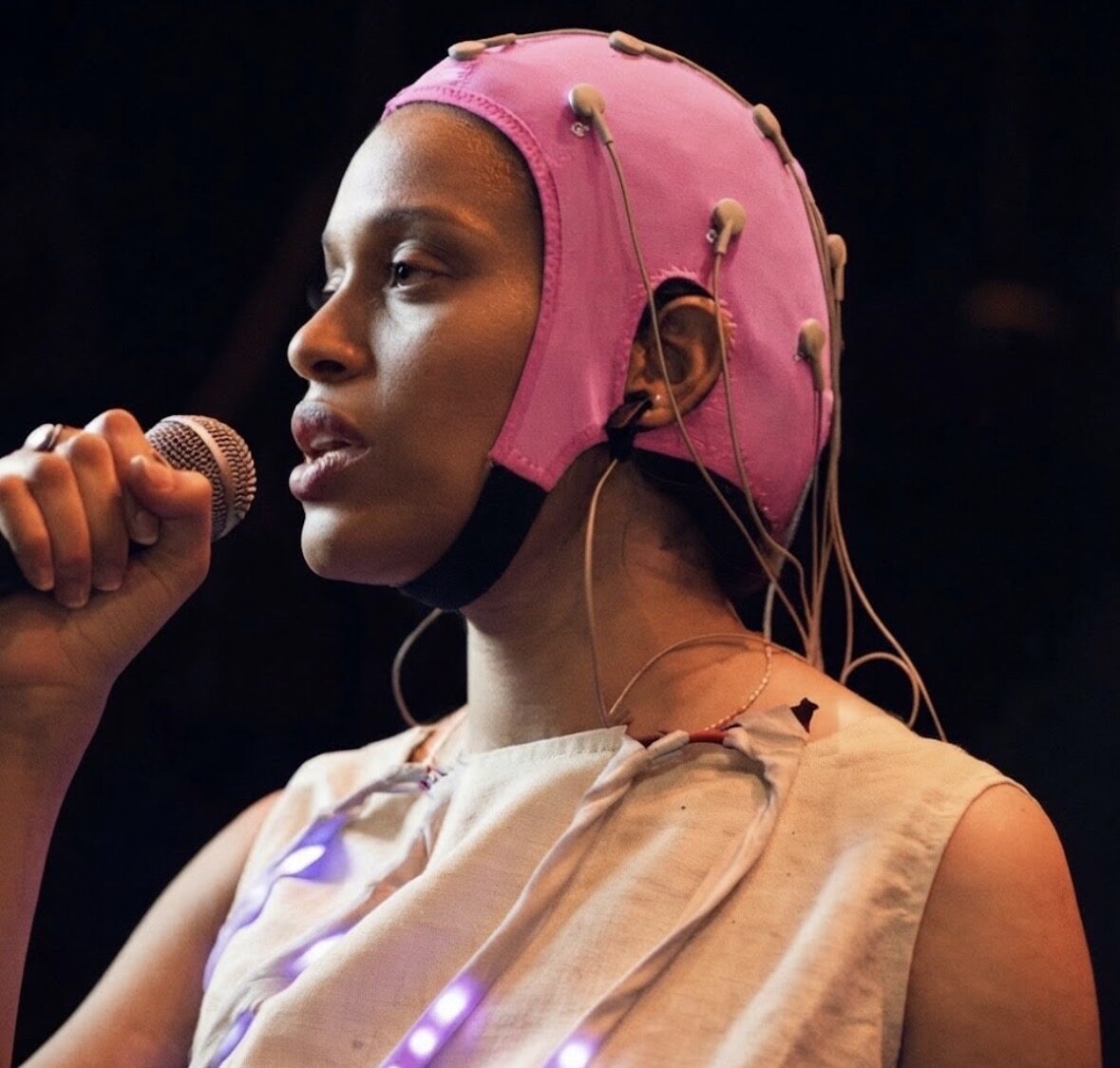 A performer in a pink EEG cap with electrode leads singing into a microphone under stage light.