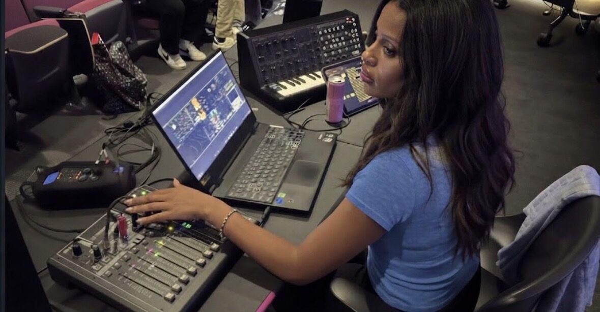 Ashlei Lewis at a mixing console with a laptop and synthesizer in a studio.
