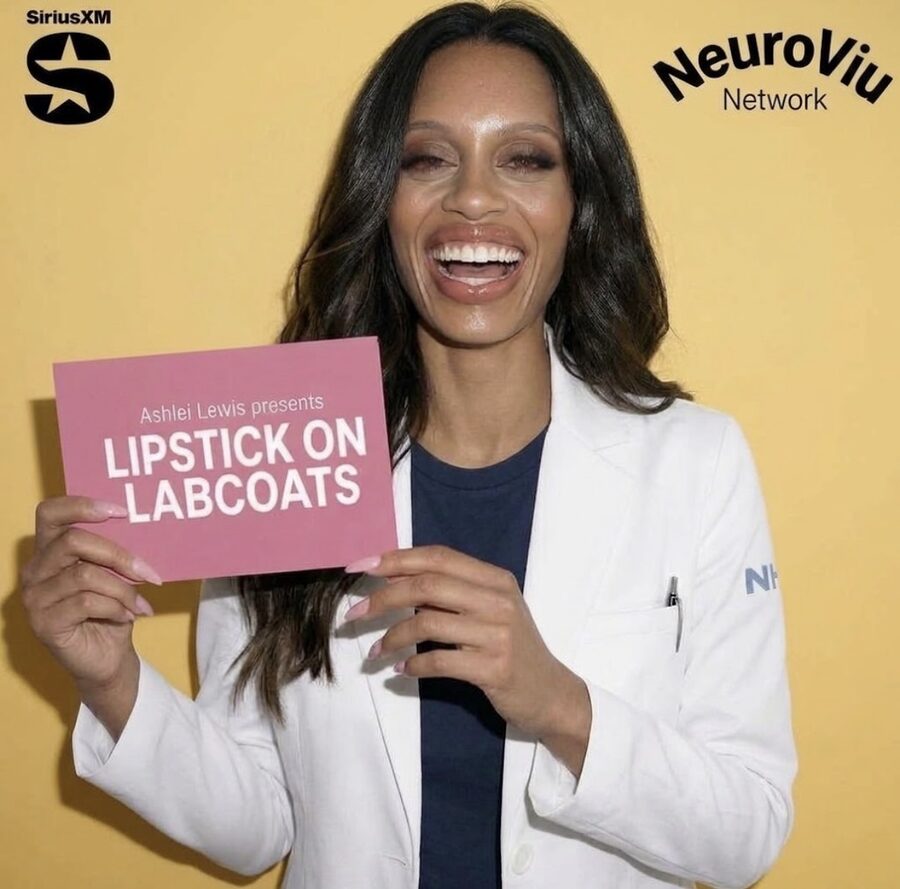 Ashlei Lewis in a white lab coat, smiling and holding a pink card that reads “Ashlei Lewis presents Lipstick on Labcoats.”